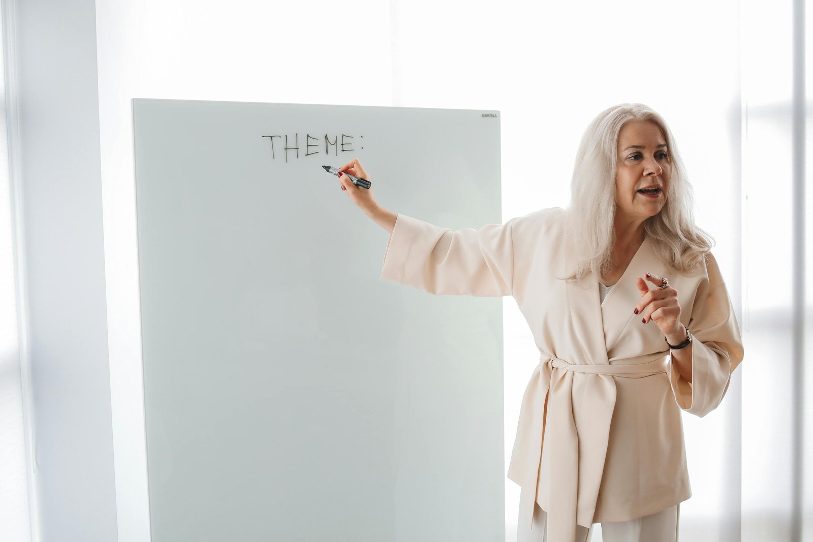 A female teacher stands at a whiteboard, with a marker pen. She has written the word, 'Theme'.