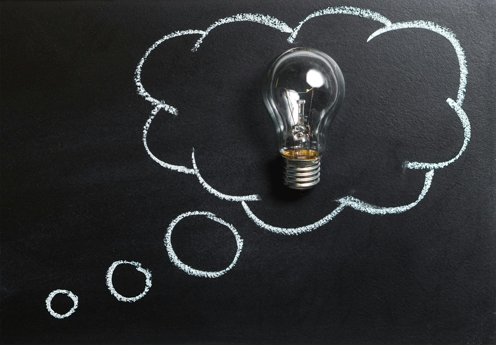 An incandescent lightbulb against a black background, with a cloud drawn around it in chalk