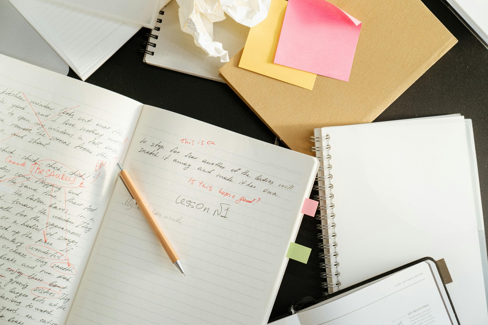 A series of notebooks scattered on a desk. One is open and has red ped corrections over the writing.