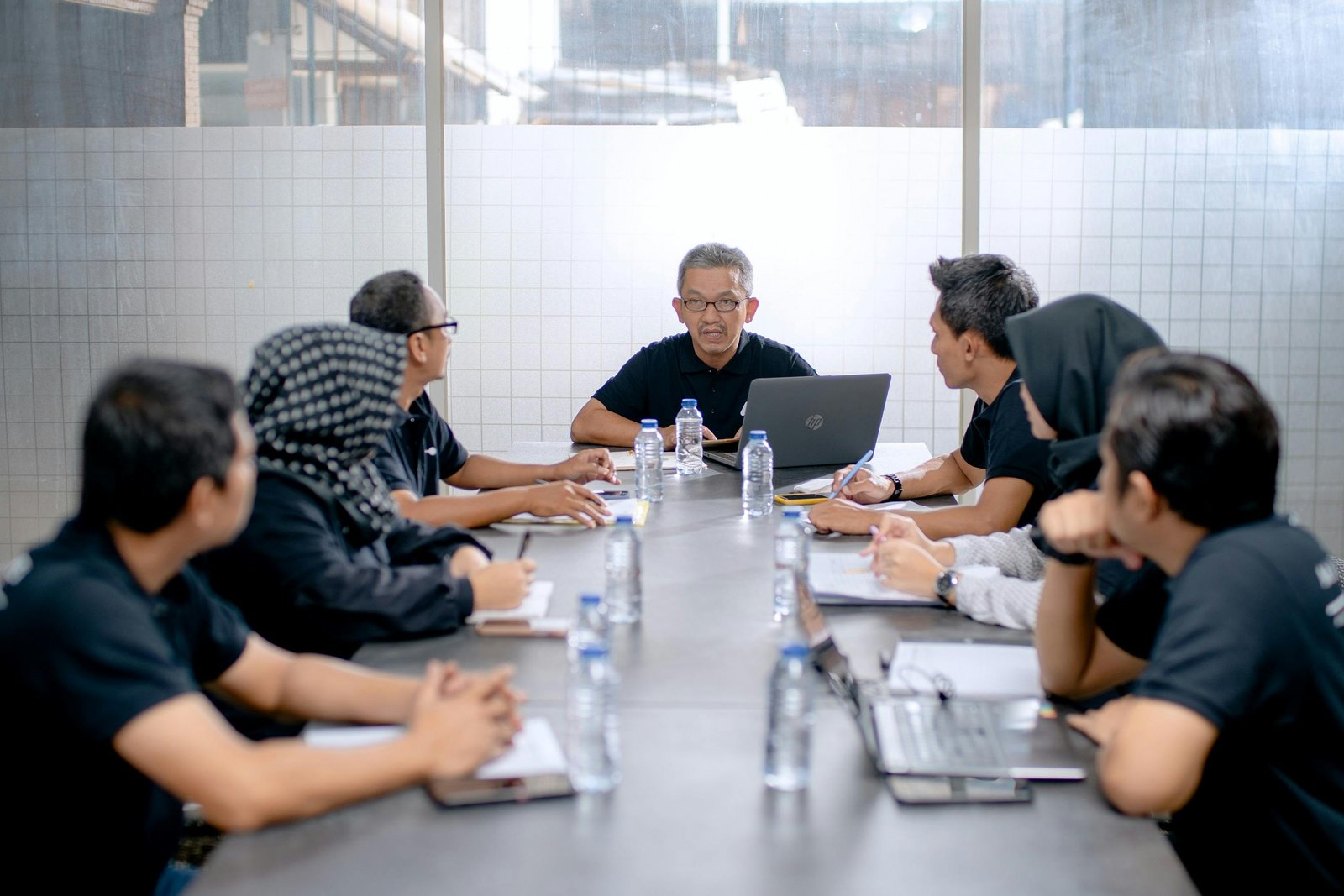 A group of employees sitting around a meeting table. They are looking at one man at the end who is speaking to them.